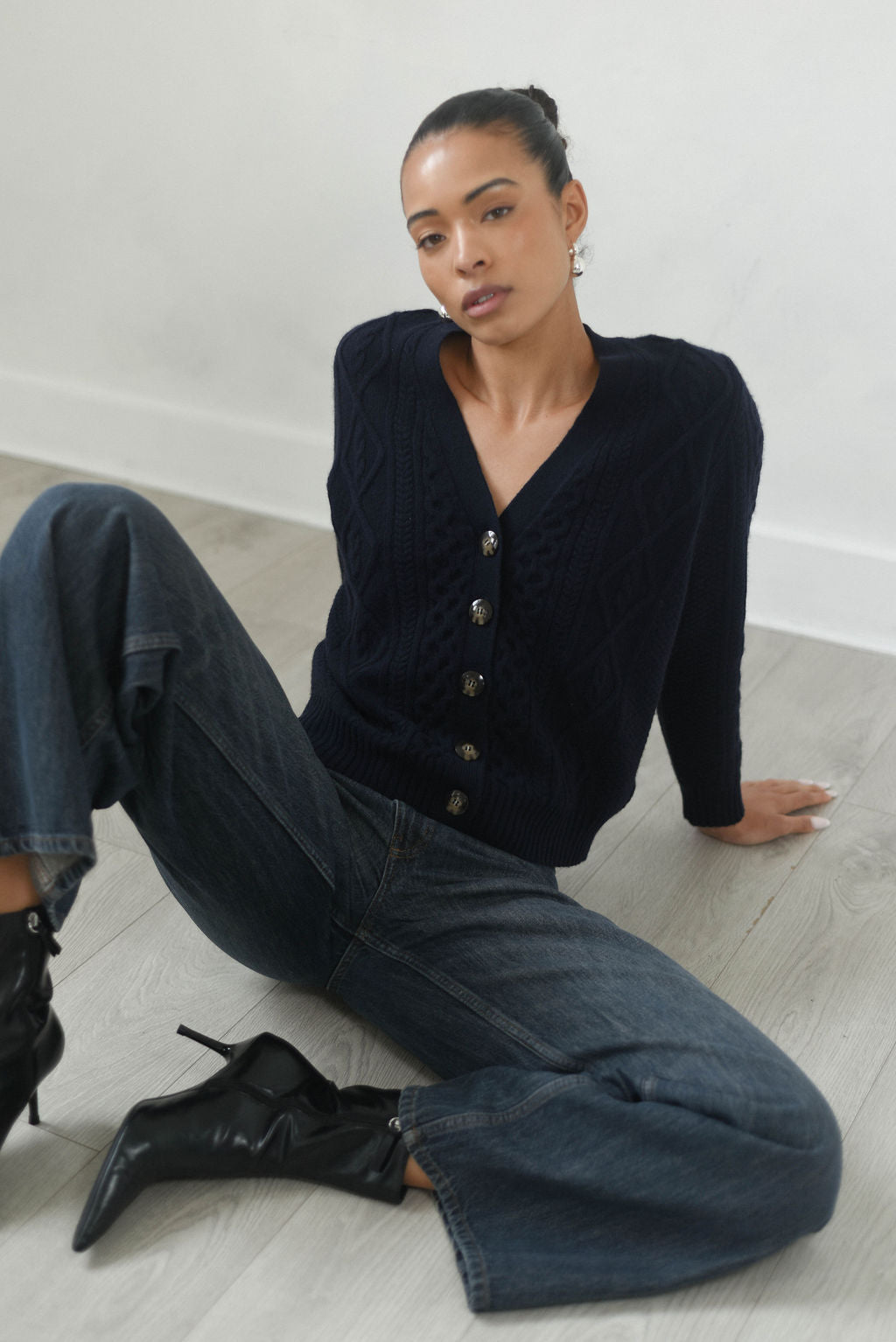 Woman sitting on a light wooden floor wearing a dark blue cardigan and jeans.